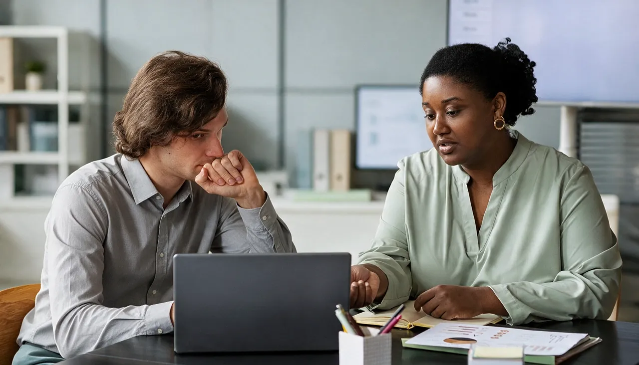 Two business professionals reviewing documents and discussing information on a laptop in a modern office