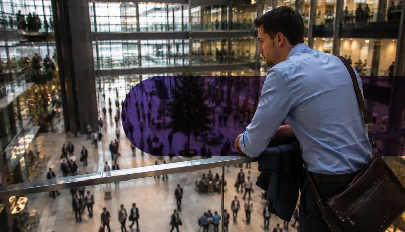 A man leans on a railing, looking down at a busy, modern atrium filled with people.