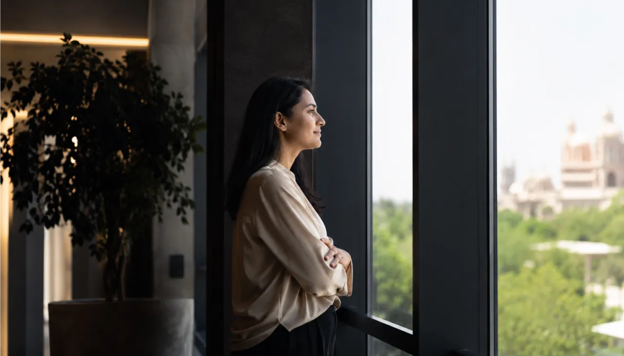 Professional woman smiles, looking out a large office window at a city skyline.