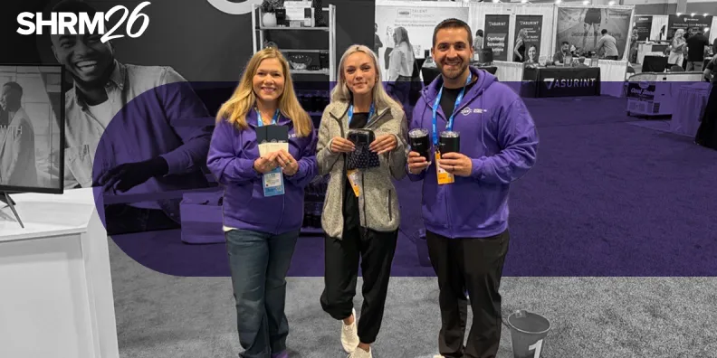 Three smiling attendees in purple hoodies holding items at the SHRM26 conference.