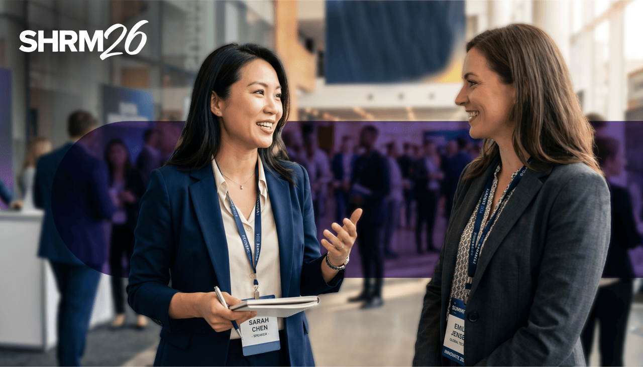 Two businesswomen converse at a SHRM26 conference, gesturing and engaging in discussion.