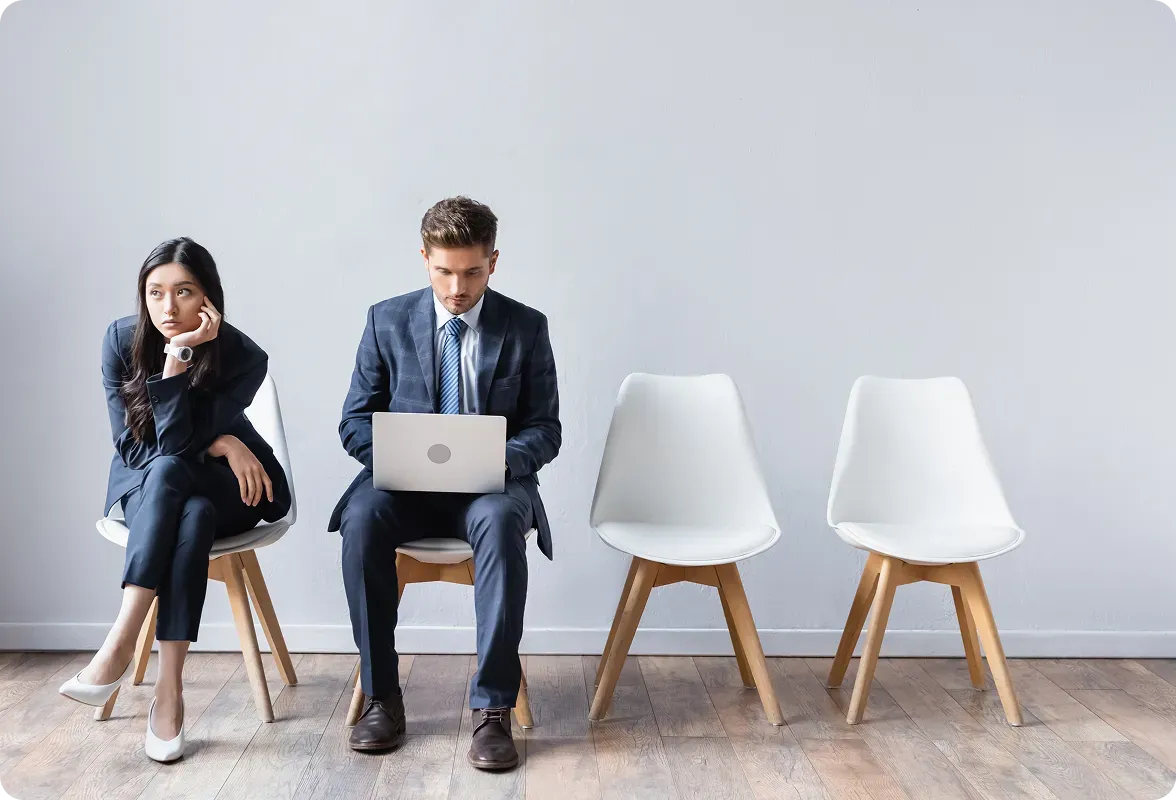 Man using a laptop and woman sitting bored in chairs in a waiting room.