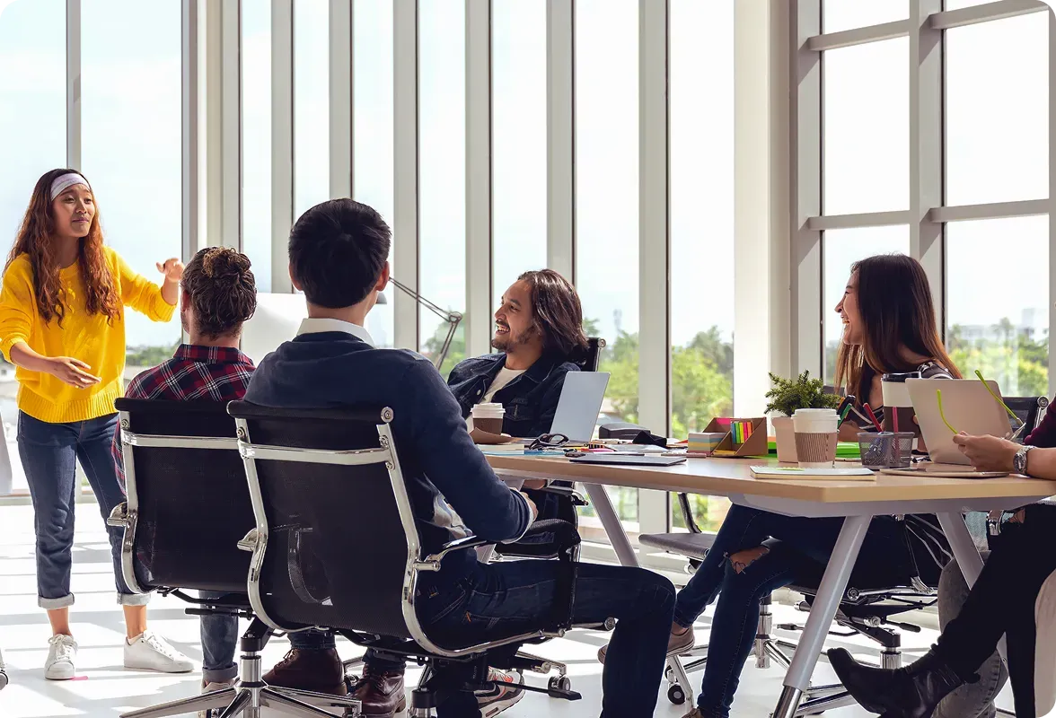 Woman in yellow shirt presenting to diverse colleagues in a bright, modern office.