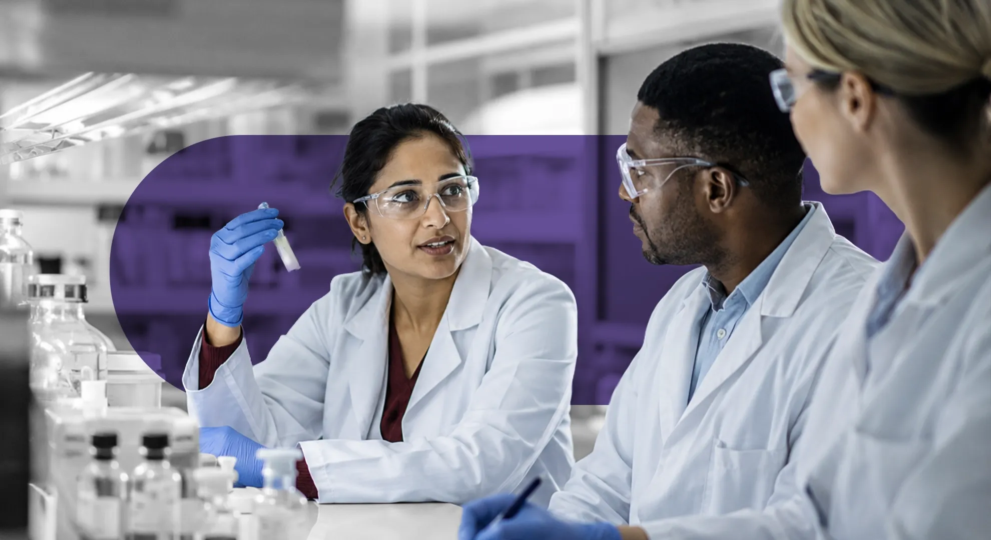 Three diverse scientists in lab coats and safety glasses discussing a sample vial in a laboratory.