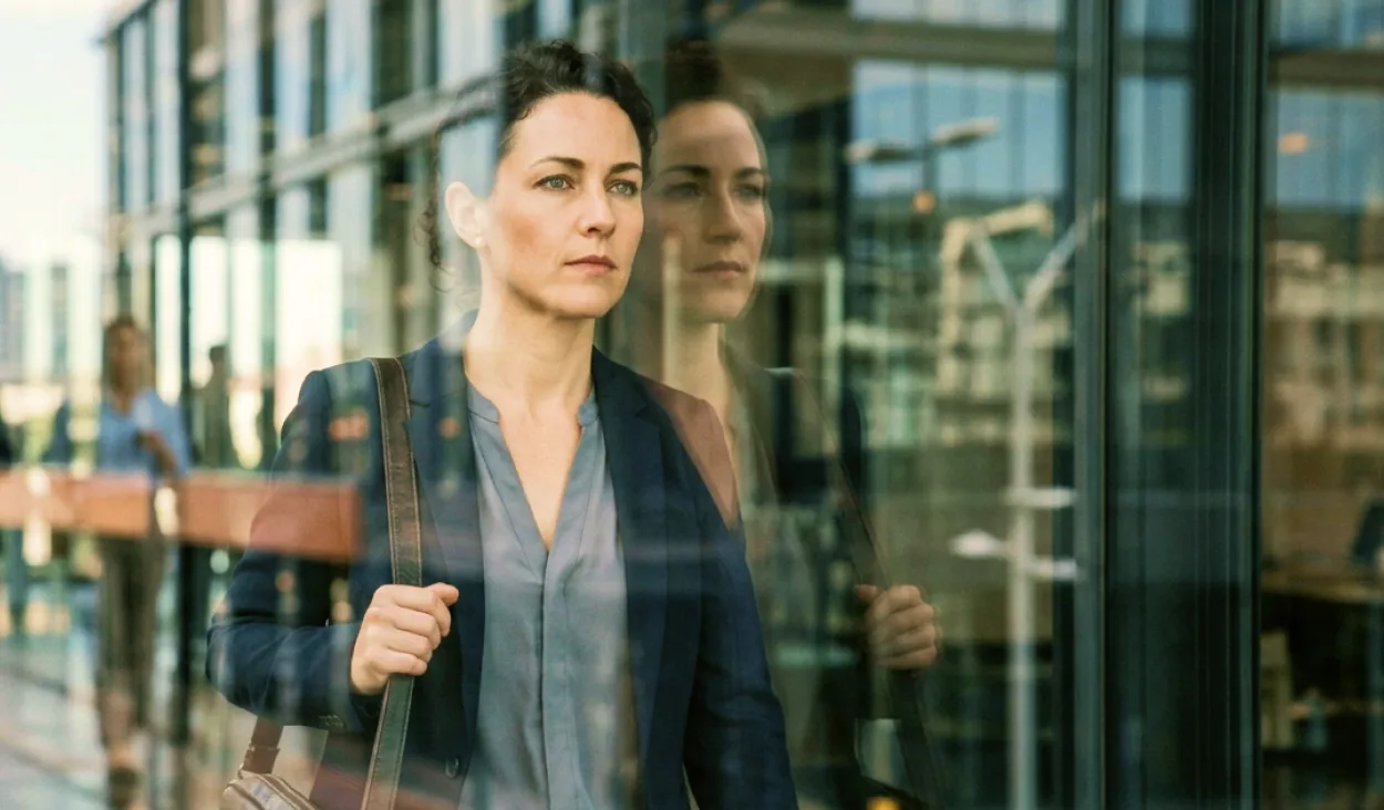 Determined businesswoman walking past a modern glass building with her reflection visible.