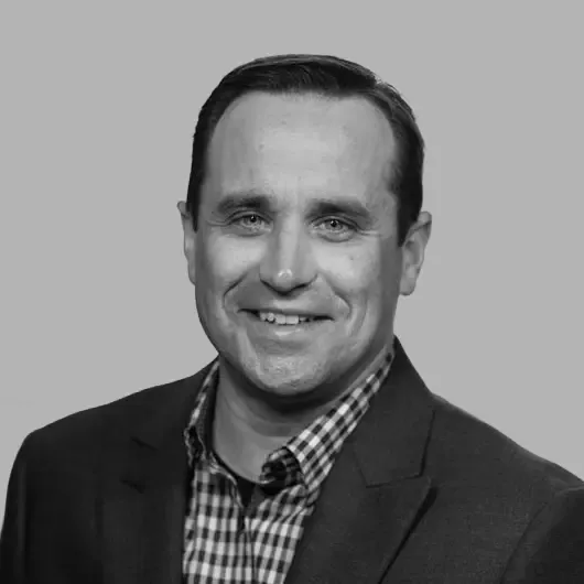 Smiling man in a suit jacket and plaid shirt against a gray background.