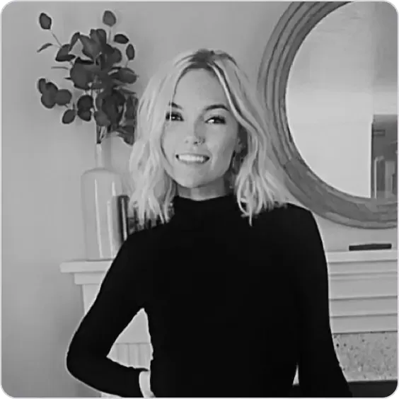 Smiling woman with blonde wavy hair wearing a black turtleneck, standing indoors.