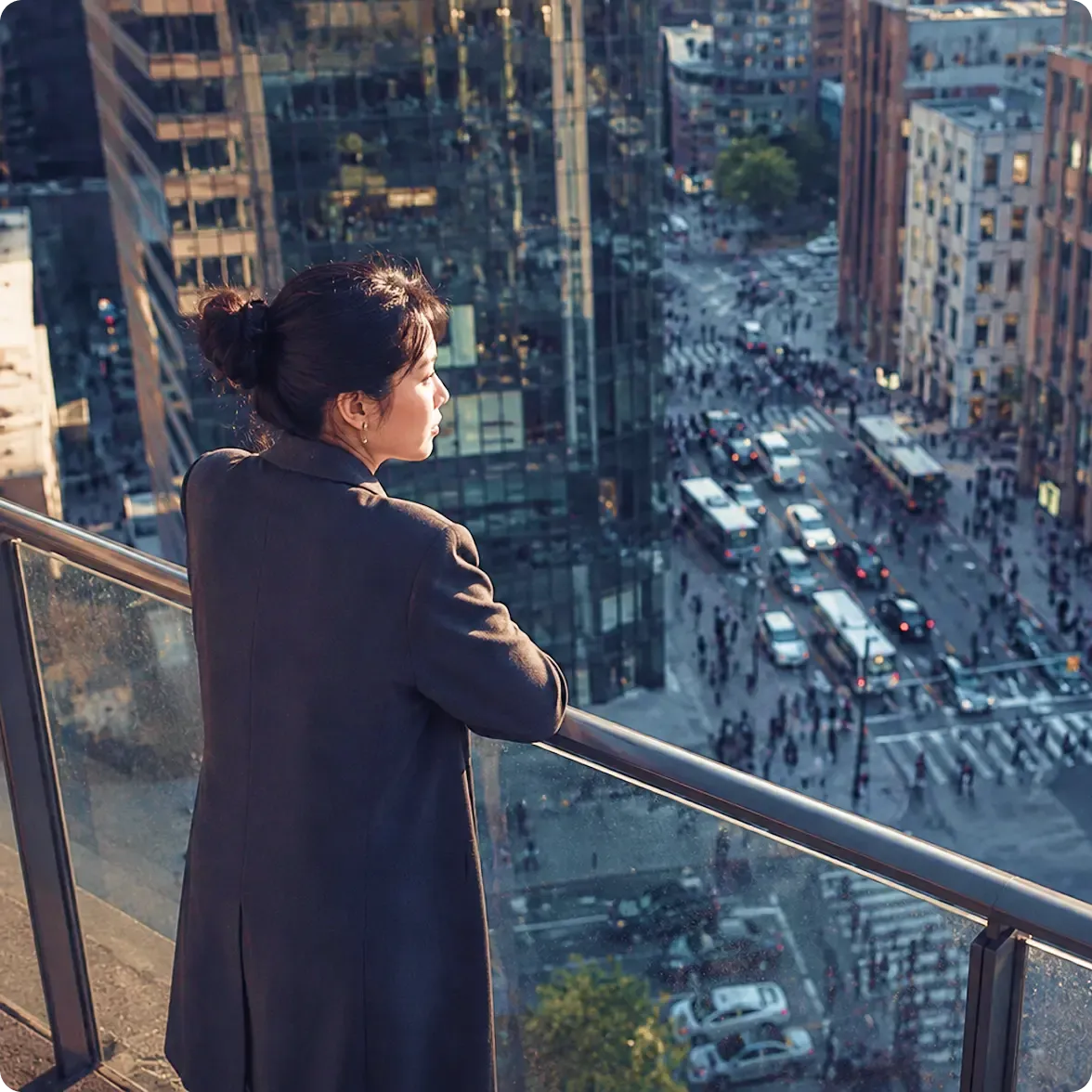 Woman on balcony overlooking a busy city street with traffic and pedestrians.