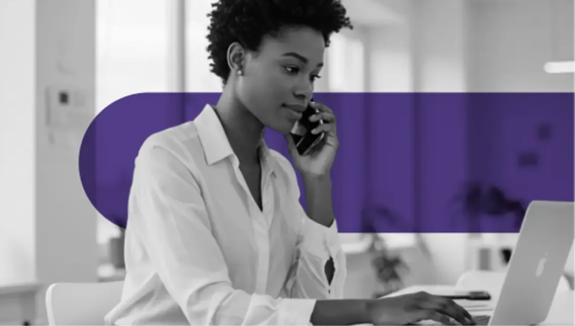 Professional Black woman talking on phone and typing on a laptop in an office.