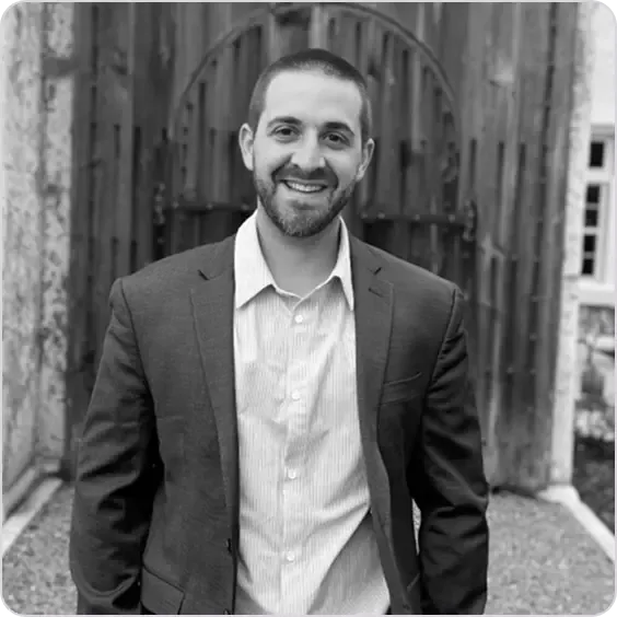 Smiling man in suit jacket and collared shirt stands before a rustic wooden door.